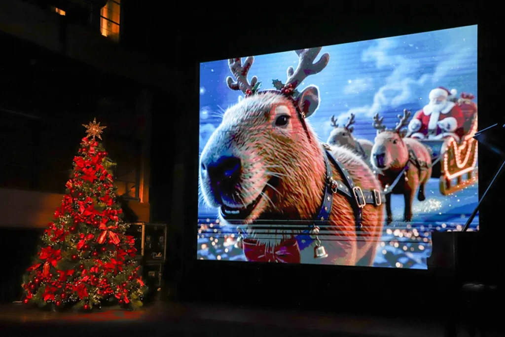Telão com as Capirrenas, mascotes do Natal de Curitiba 2026 que misturam capivara e rena, ao lado de uma árvore de Natal iluminada.