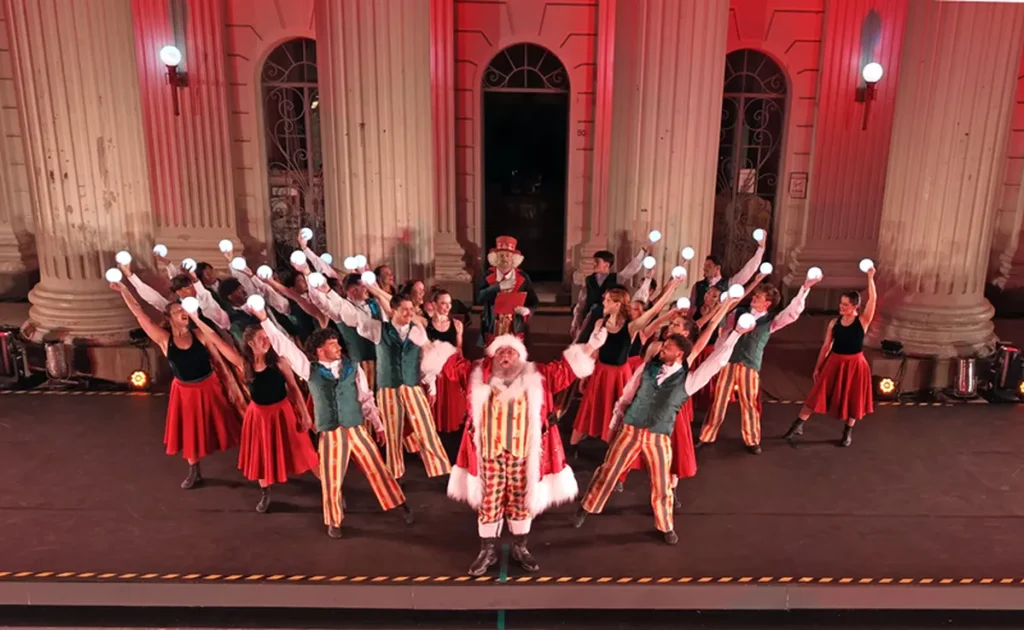 Elenco com figurinos natalinos e Papai Noel se apresenta em palco em frente a um prédio histórico iluminado de vermelho, em Curitiba.